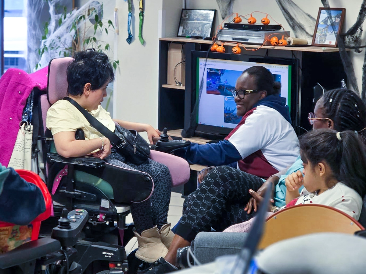 Une femme en fauteuil roulant interagit avec une animatrice devant un écran affichant un environnement virtuel, entourées d'autres participants dans une salle décorée.