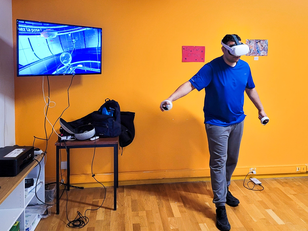 Un homme en tee-shirt bleu porte un casque VR et tient des manettes devant un mur orange vif, avec un écran à gauche montrant son point de vue dans l'environnement virtuel.